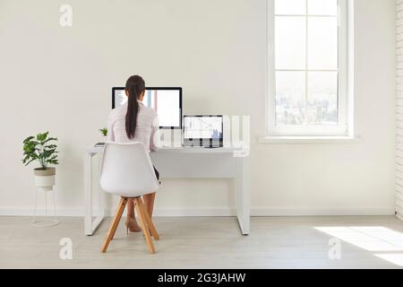 Back view of woman sitting at office desk and working on desktop computer and laptop Stock Photo