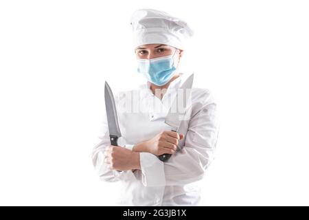 Isolated chef with mask due to pandemic standing in a white cooking robe with crossed hands holding two sharp knives. Stock Photo