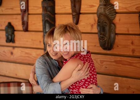 Two women 55 years old girlfriends hugging against the background of wooden wall, posing for photographer Stock Photo