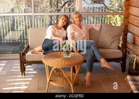 Lovely ladies 55 years old are bothered on sofa at table with flowers in an authentic setting of a wooden house Stock Photo