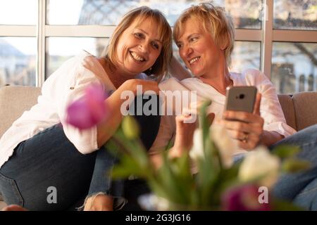 Cheerful summer evening of two girlfriends 55 years old on veranda of wooden house, use smartphone Stock Photo