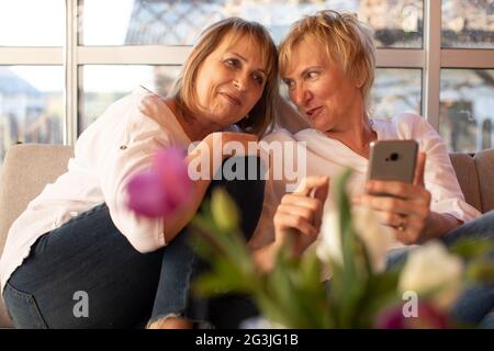 Cheerful summer evening of two girlfriends 55 years old on veranda of wooden house, use smartphone, confidential conversation Stock Photo
