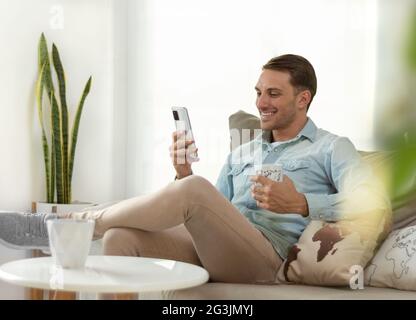 young man smiling, holding and looking at his smartphone, relaxed on the sofa at home drinking a coffee Stock Photo