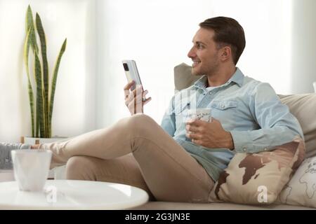 handsome young man, holding his smartphone, looking out the window, making a video call, while having coffee relaxing on the sofa at home Stock Photo