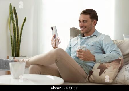 handsome young man, holding his smartphone, making a video call, while having coffee relaxing on the sofa at home Stock Photo