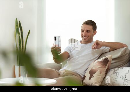 young man holding a smartphone, making a video call, waving at the screen. sitting on the sofa at home relaxed Stock Photo