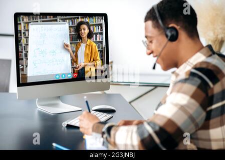 Distant learning, webinar, online lecture. Hispanic clever male student, sits at work desk, uses computer, listening to an online lecture, taking notes, female african american teacher on the screen Stock Photo