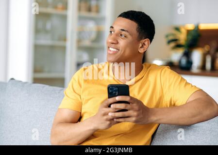 Pensive happy young latino man in a yellow casual t-shirt, sitting at home on the couch, holding his smartphone in his hand, texting online, browsing social networks, looking away, smiling Stock Photo