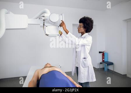 Young confident Afro American female doctor radiologist, taking x-ray scan of young male patient lying on the machine table. Doctor standing near the patient during chest X Ray procedure Stock Photo