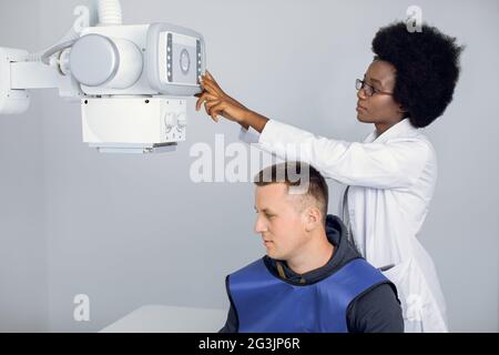 Young afro american female radiologist technician, setting up the modern machine to take x-ray scan, while performing scanning of male patient broken hand. Radiologist and patient in a x-ray room Stock Photo
