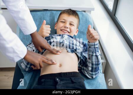 Top angle view of young female African Amrican unrecognizable doctor palpating belly of little boy lying on couch in doctors office and smiling showing thmbs up to camera Stock Photo