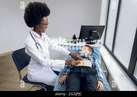 Portrait of smiling joyful African lady doctor performing manual palpation, examining stomach abdomen of little boy and talking to him where it hurts. Belly pain concept Stock Photo