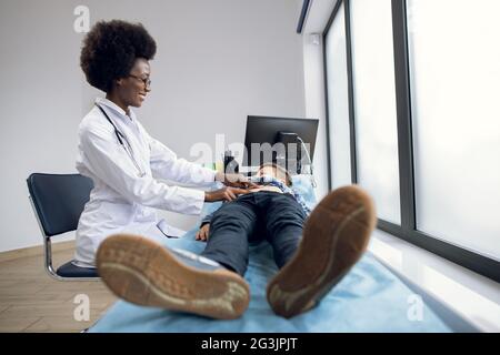 Female African American doctor gp pediatrician, in white coat palpating belly of teen child boy, lying on couch, smiling and talking to little patient. Abdominal pain, medical annual checkup concept Stock Photo