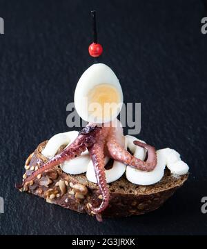 Creative and unusual sandwich - Spanish tapas similar to a sea monster, consisting of octopus tentacles and a cut quail egg on cream cheese Stock Photo
