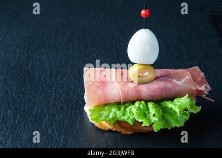 Creative Spanish tapas sandwich consisting of quail egg, green olive,  and Spanish jamon - ham on a leaf of green lettuce on a baguette bruschetta - s Stock Photo