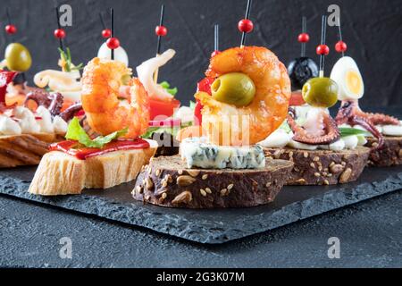 Traditional Spanish food - tapas of a large fried shrimp on gorgonzola cheese with a green olive in the middle on a black baguette with sesame seeds - Stock Photo