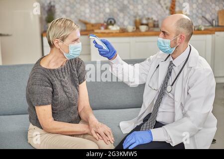 Man doctor measures the temperature of an elderly woman at her home wearing a protective mask. Coronovirus infection diagnosis concept Stock Photo