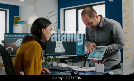 Industrial designer discussing with colleague holding tablet with in CAD program, designing 3D prototype of building. Woman engineer using pc with two monitors sharing ideas for construction mechanism Stock Photo