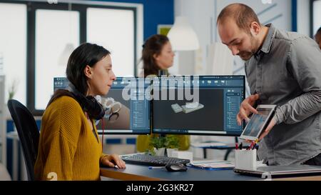Industrial designer holding tablet discussing with woman engineer while working in CAD program, designing new components, comparing 3D prototype with project from notepad, using pc with two monitors Stock Photo