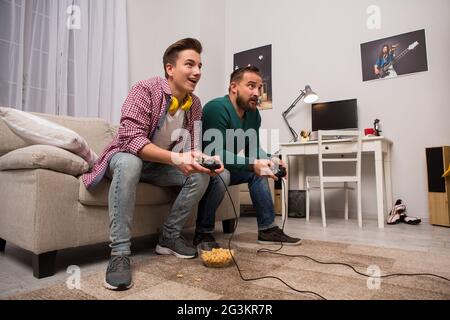 Side view of father and teenage son sitting on sofa, playing video games.