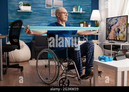 Disabled senior man in wheelchair training with elastic band exercising body workout recovering after disability accident watching strech video on tablet. Pensioner doing healthcare arm exercise Stock Photo