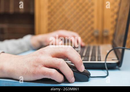 The hands of an unknown person using a laptop, moving the cursor with the mouse and typing with the keyboard, on a blue table. Stock Photo
