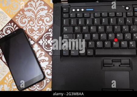 Part of the keyboard of a laptop and a smartphone can be seen with some reflection on its screen on a table with a decorative mat. Stock Photo