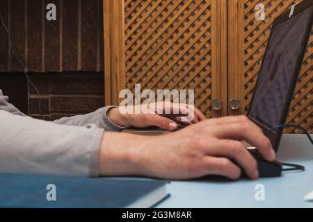 The hands of an unknown person using a laptop, moving the cursor with the mouse and typing with the keyboard, on a blue table. Stock Photo