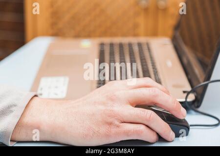 The hands of an unknown person using a laptop, moving the cursor with the mouse and typing with the keyboard, on a blue table. Stock Photo