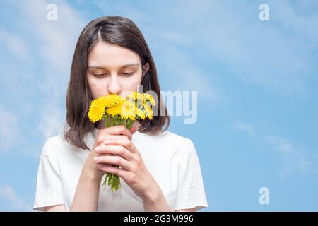 Pretty caucasian girl inhales the scent of yellow dandelions against a blue sky with clouds, copy space. The girl received a bouquet of wildflowers as Stock Photo