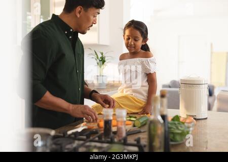 Happy hispanic daughter and father preparing vegetables in kitchen, daughter sitting on counter Stock Photo