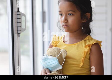 Sad hispanic girl standing at window looking out of window holding teddy bear in face mask Stock Photo