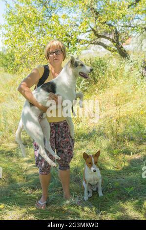 Nice outdoor portrait of mature Ukrainian woman taking on the hands big white  mixed breed dog while basenji one sitting next in apricot orchard at su Stock Photo