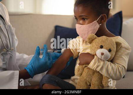 African american female doctor giving covid vaccination to girl patient in face mask at home Stock Photo