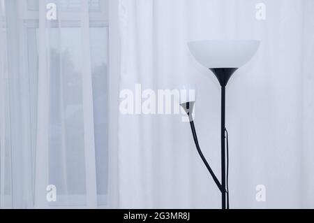 Floor electric lamp in black with white lampshades in the interior of the room against the background of white curtains and windows. Stock Photo