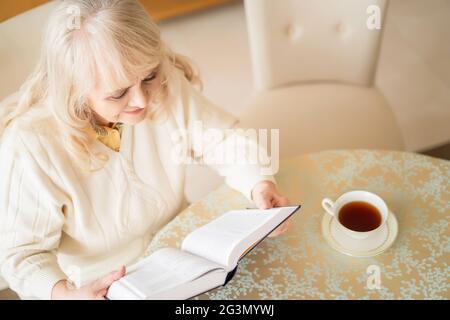 Elegant senior woman reads book at table and having tea Stock Photo