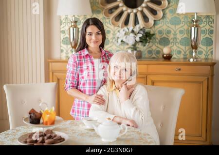 Beautiful daughter gently hugs her senior mother who sits at the table Stock Photo