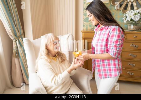 Charming daughter gives small birthday cupcake with candle to her senior mother Stock Photo