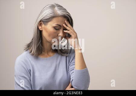 Mature Asian woman touches her forehead in distress Stock Photo