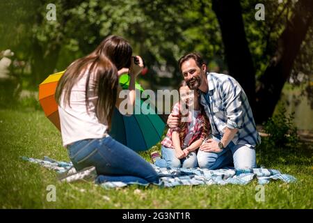 Young Mother Making Photo Of Husband And Daughter In The Park. Father Is Hugging His Daughter While Sitting On The Blanket. Colo Stock Photo