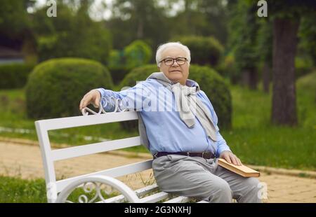 Senior man sits on a bench and reads a book. An elderly man with White hair is resting on a park bench Stock Photo