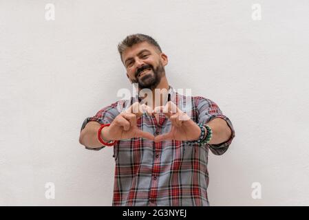 Beautiful caucasian man making heart sign to the camera on white wall background.Love concept. Stock Photo