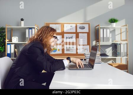 Side view of a funny strange male office worker working on laptop sitting at desk in office. Stock Photo
