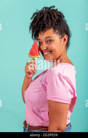 Black girl eating a slice of watermelon in the form of ice cream on a turquoise background Stock Photo