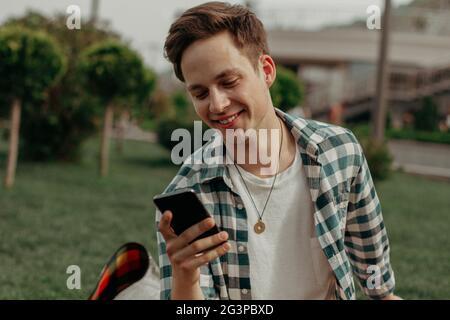 Cute European Guy Smiles Message Looking Into Mobile Sitting On Grass Stock Photo