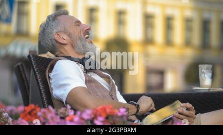 Grey bearded man laughs from the news on a tablet on a sunny day in a cafe Stock Photo