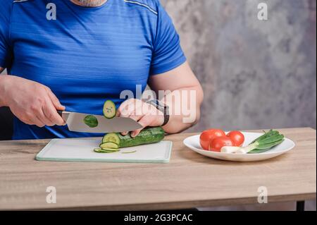 A man cuts a cucumber on a board. Tomato and green onions lie on a white plate Stock Photo