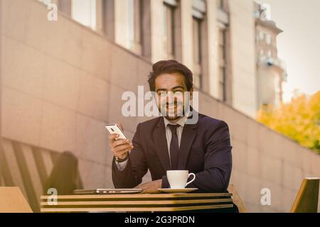 Businessman using mobile phone outdoors. Successful man toothy smiles holding smartphone while sitting at table with cup of coff Stock Photo