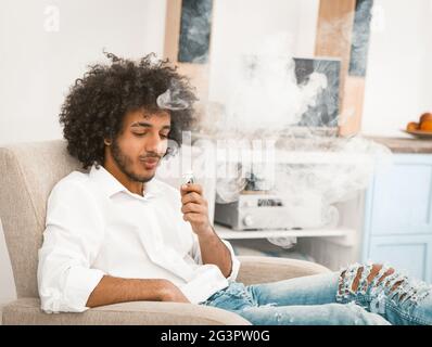 Bearded man enjoying puffs of smoke of electronic cigarette. Young man in casual rests at home sitting in an armchair and smokin Stock Photo