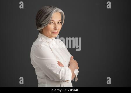 Pretty white-haired woman smiles looking at camera with arms crossed. Stylish caucasian businesswoman isolated on dark gray back Stock Photo